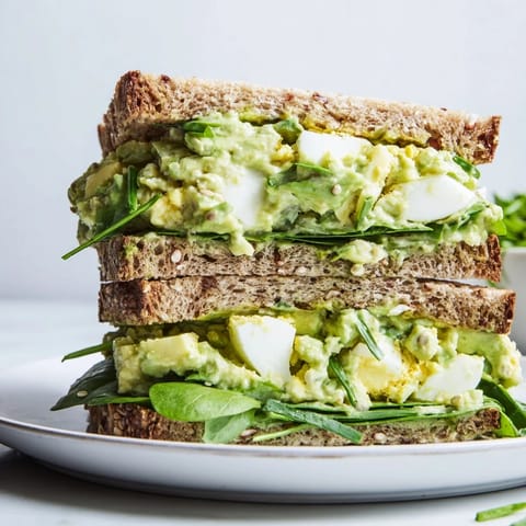 Close-up view of a homemade avocado egg salad sandwich, highlighting the chunky texture of the eggs mixed with smooth avocado and bright chives on toasted bread.