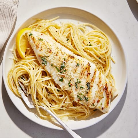 A close-up of Lemon Bass Pasta showcases flaky sea bass, a zesty garlic-lemon glaze, and a sprinkle of Parmesan cheese.