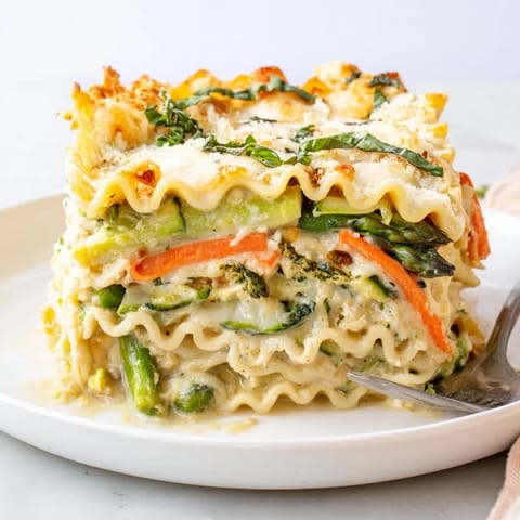 A close-up slice of Spring Veggie Alfredo Lasagna reveals creamy ricotta, melted mozzarella, and vibrant spring vegetables like carrots and snap peas between no-boil noodles.
